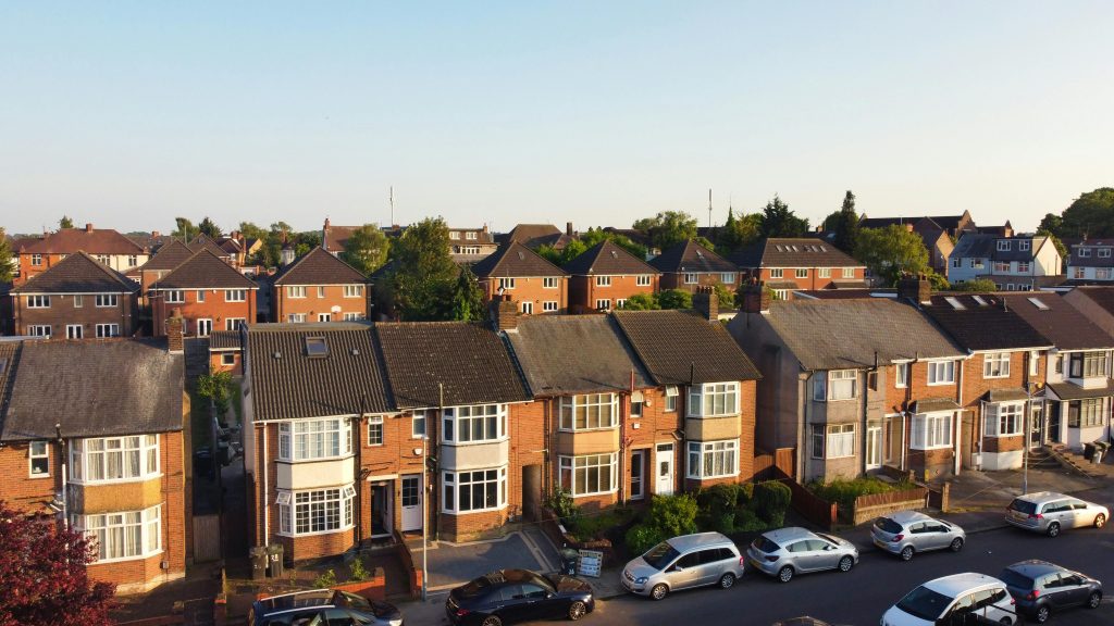 Suburban street with brick houses and cars parked, basking in warm sunlight, surrounded by greenery.